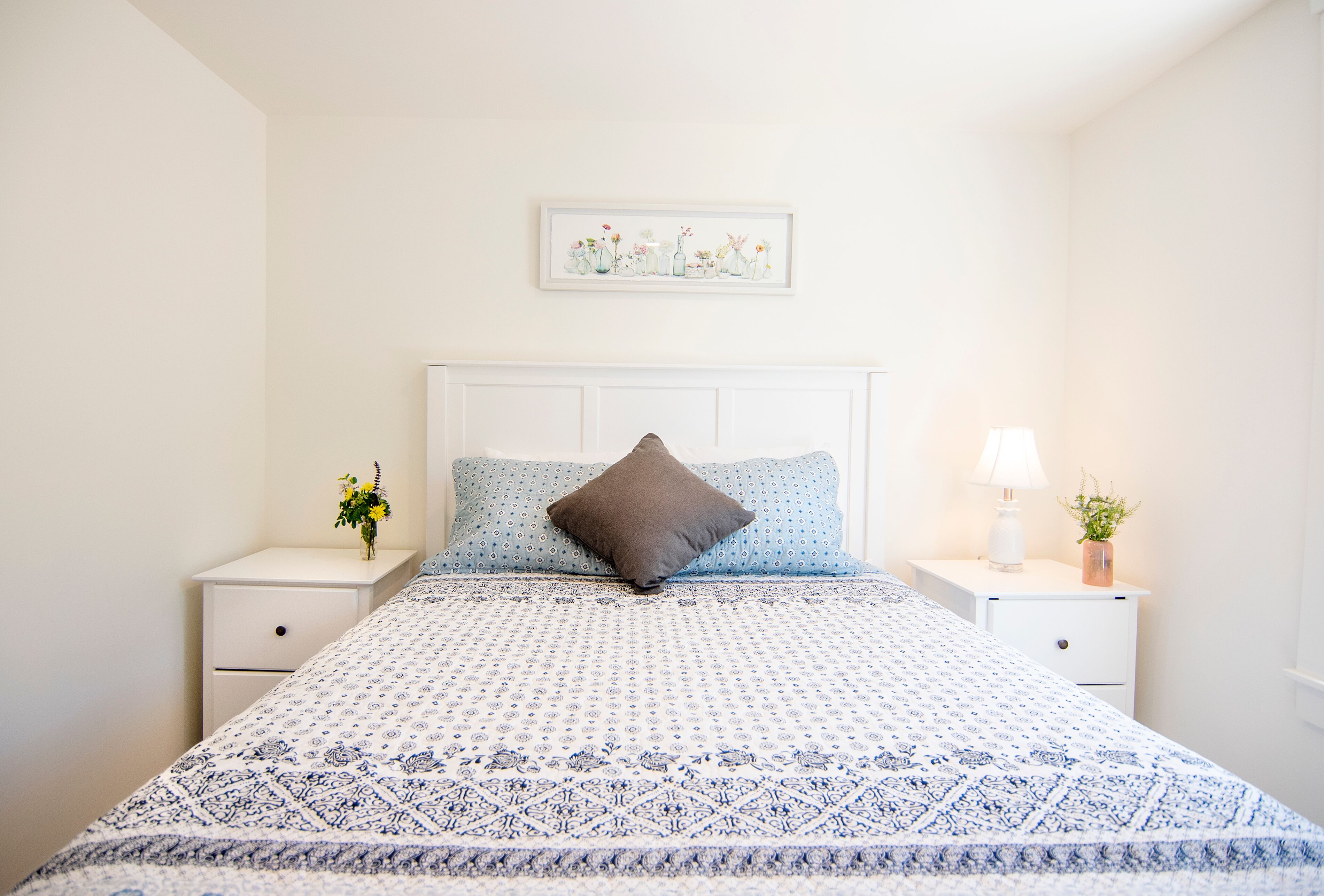 Cama bem arrumada com colcha estampada, um travesseiro e um quadro emoldurado acima. Duas mesas de cabeceira, cada uma com uma luminária e uma pequena planta.