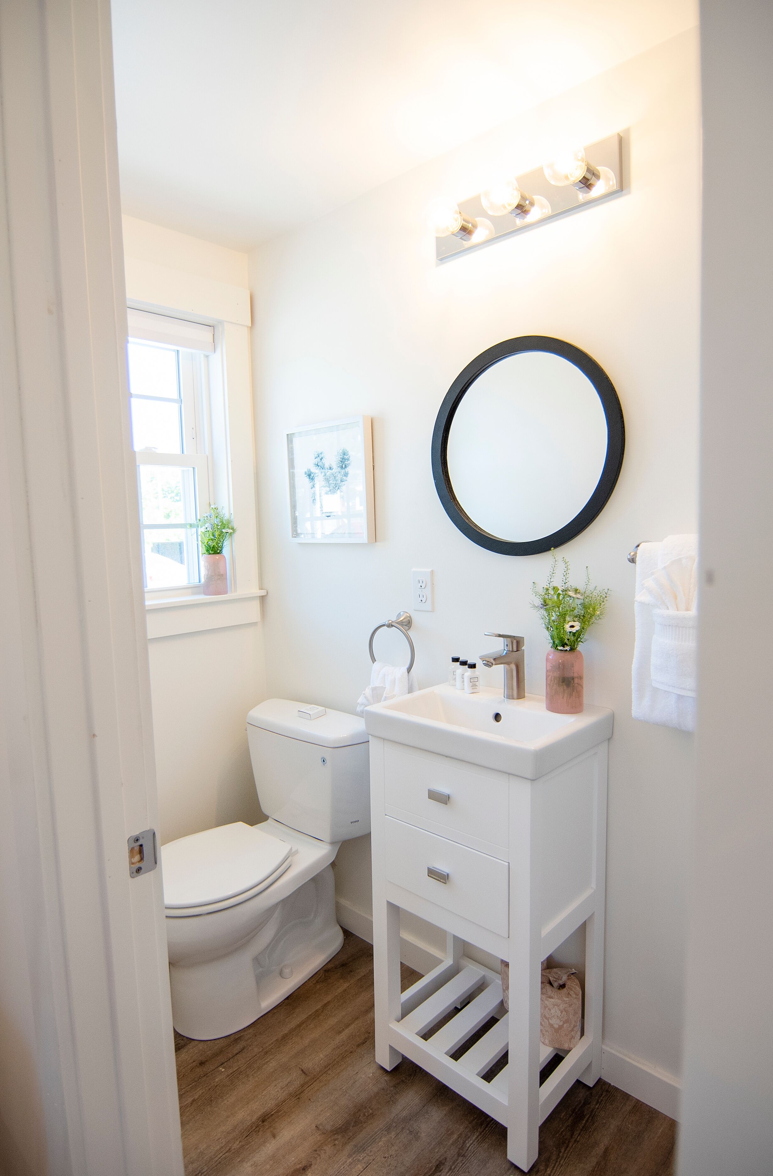 A bathroom with a white sink, mirror, and toilet. There is a window with a plant on the windowsill and a towel hanging on the wall.