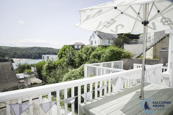 Eine Holzdielen-Terrasse mit weißen Geländern, die auf eine Küstenlandschaft mit Strand und Hügeln blickt.