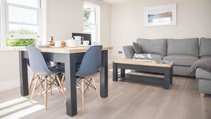 A modern living room with a grey sofa, a dining table with chairs, and a coffee table. There are windows providing natural light and a framed picture on the wall.