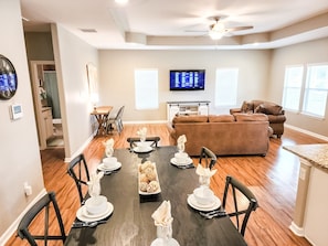 A dining area with a dark wooden table set for four, surrounded by black chairs. A brown leather sofa and a television are visible in the background. - Canyon Lake Oasis! 3 MIN to Boat Ramp Pet Friendly (Canyon Lake)