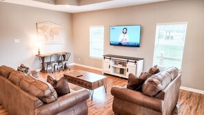 Een woonkamer met twee bruine leren banken, een houten salontafel, een televisie aan de muur, een kaart aan de muur, en een zithoek met een tafel en stoelen.
