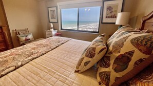 A bedroom with a bed, a nightstand, a lamp, and a window with a view of the beach.