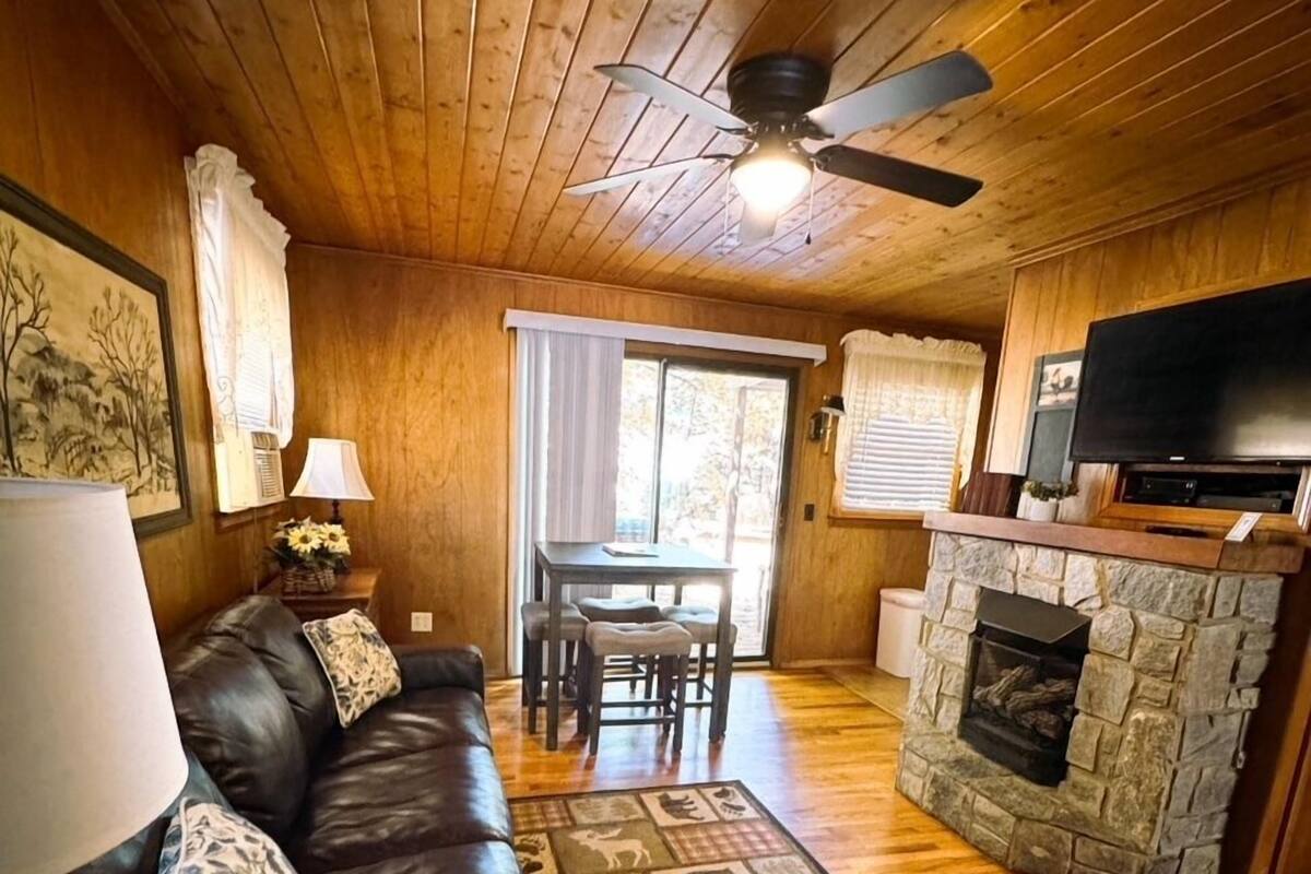 Interior acogedor de una cabaña con sofá de cuero, chimenea de piedra, televisor y ventilador de techo.