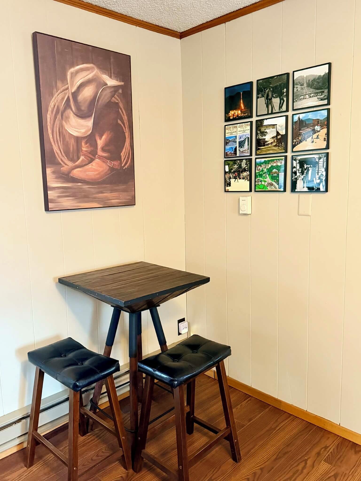 A corner room with a wooden table and two chairs, a large painting on the wall, and a series of framed photographs.