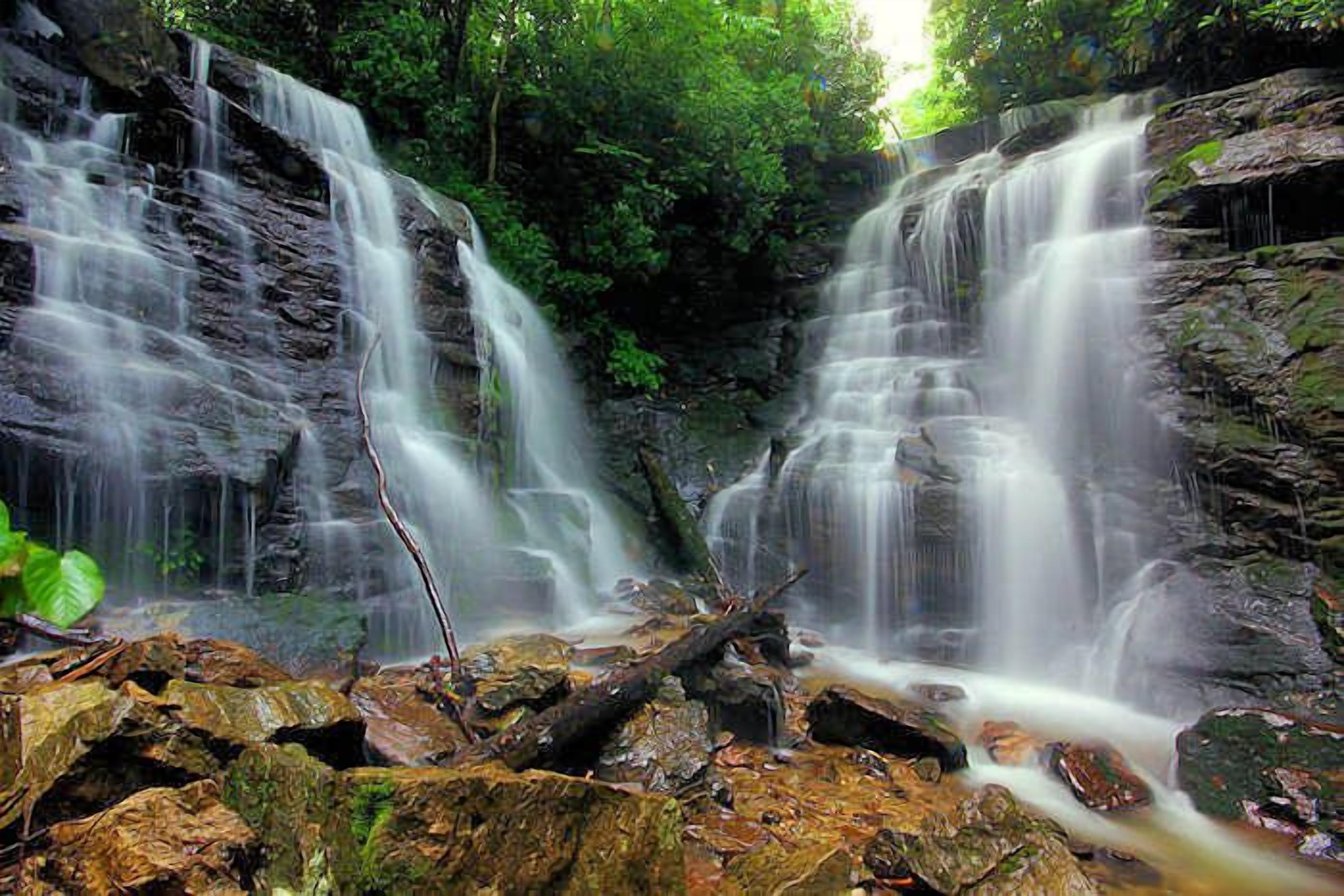 Ein Wasserfall stürzt über felsiges Gelände, im Hintergrund üppiges Grün.