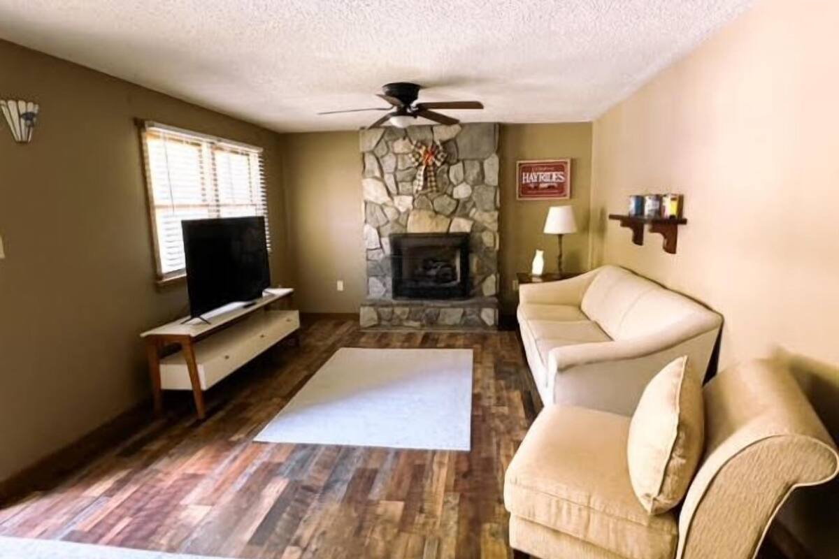 Una sala de estar con chimenea de piedra, ventilador de techo, televisor y sofá color beige.