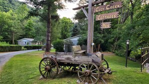Un letrero de madera rústico indica 'Cozy Creek Cottages' y da indicaciones hacia 'Yes', junto con un carruaje de madera antiguo, todo en un entorno verde y exuberante.