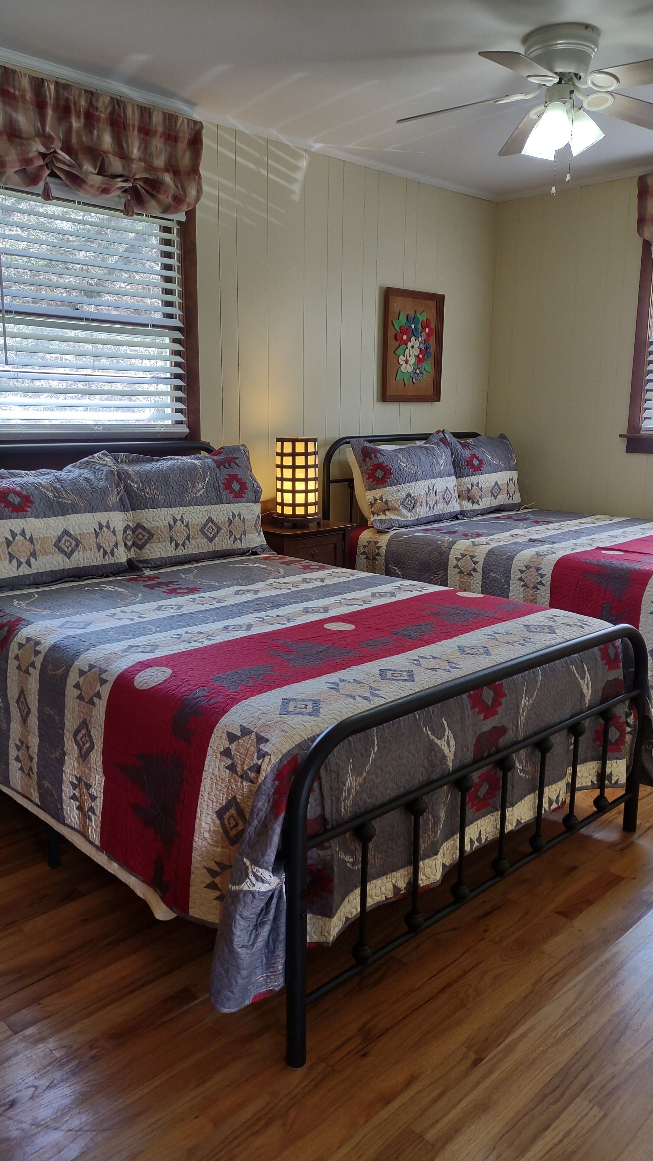 A bedroom with two beds, a ceiling fan, a window with blinds, a wall-mounted lamp, and a floral painting.