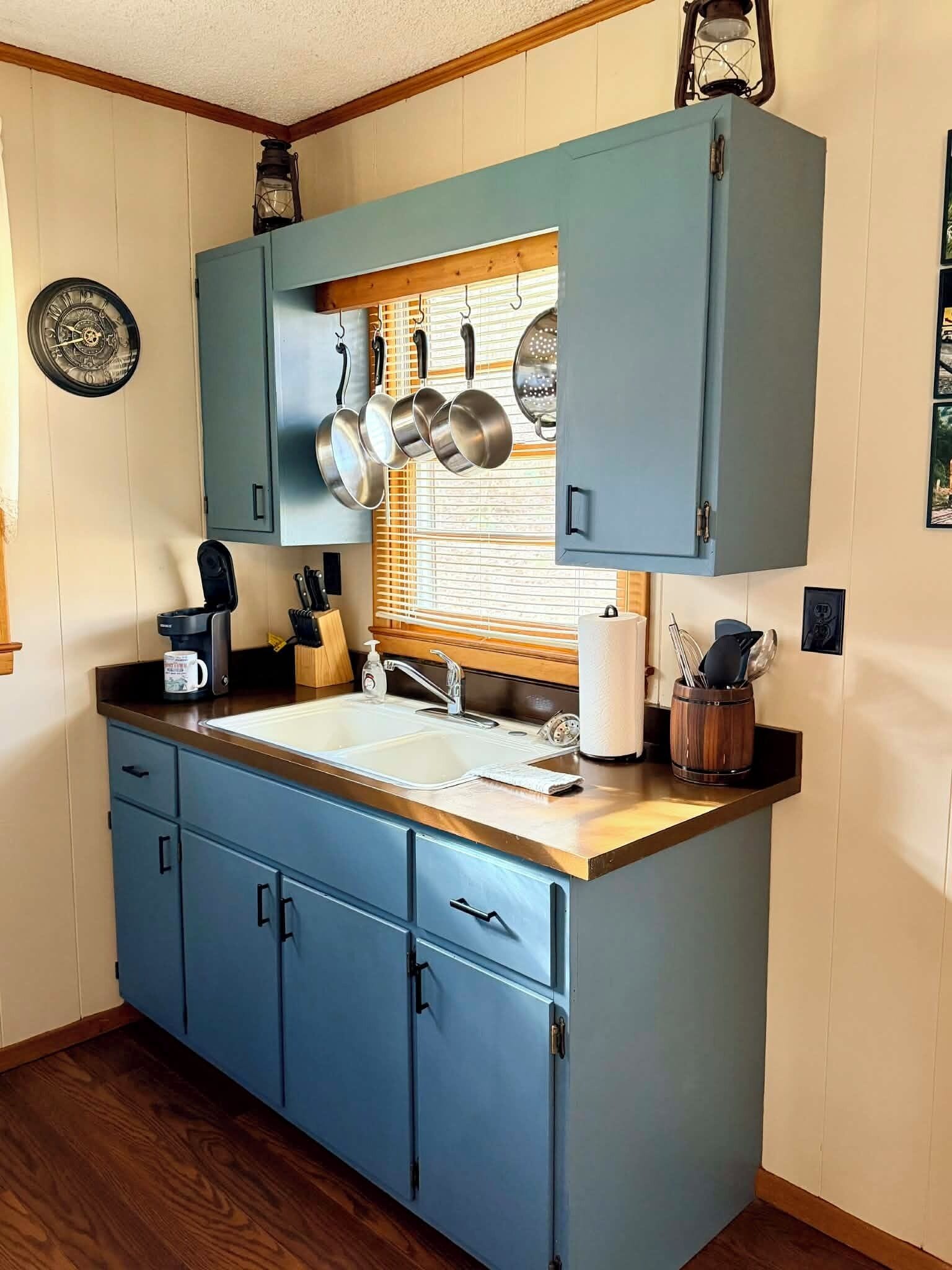 Una cocina con gabinetes azules, encimera de madera, una ventana con persianas y ollas colgando.
