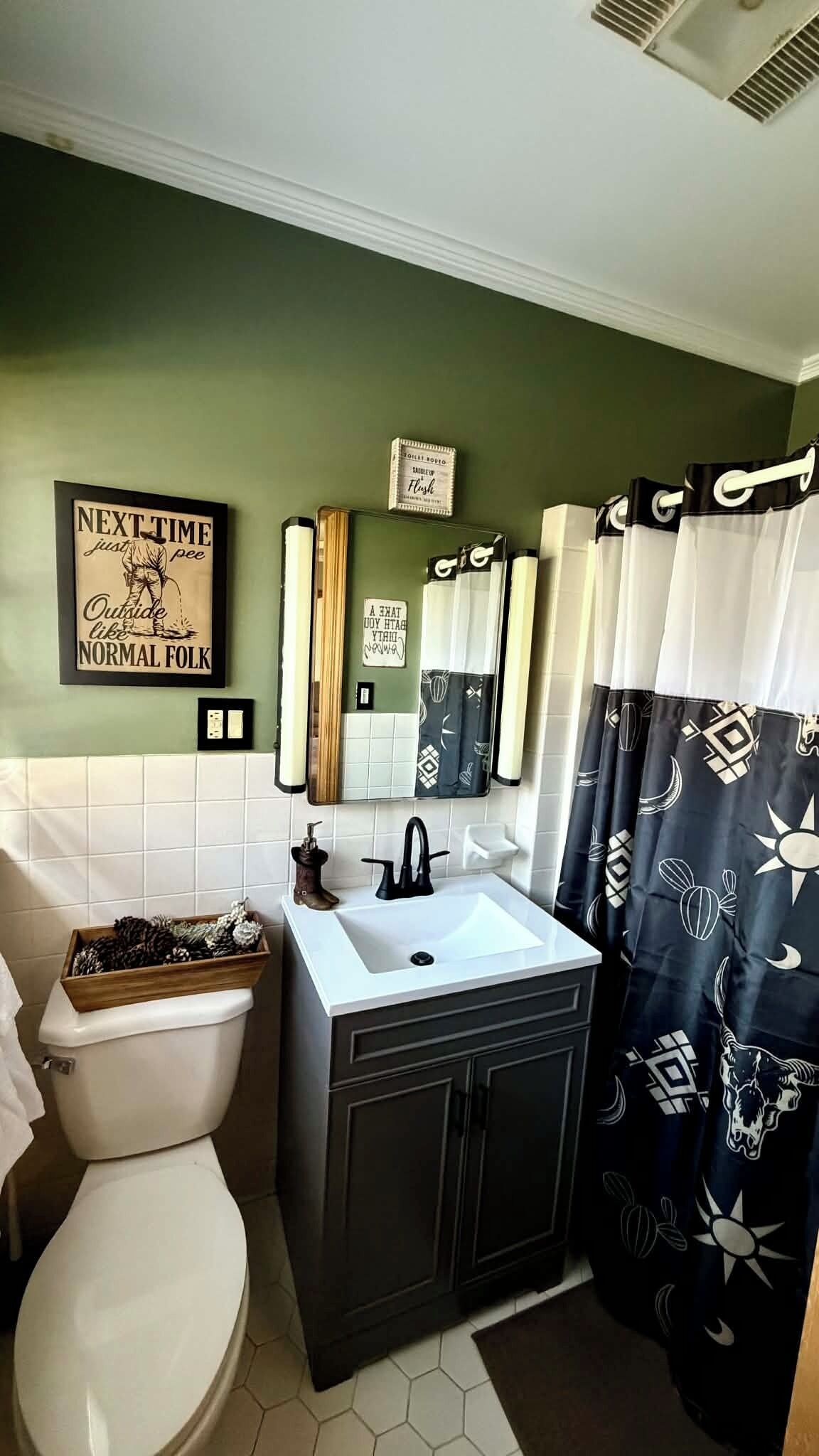 A bathroom with a toilet, sink, and mirror. There are decorative items on the toilet and a mirror with a sign above it.