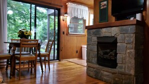 Un interior de madera con chimenea de piedra, una zona de comedor con sillas y mesa, y una ventana con persianas.