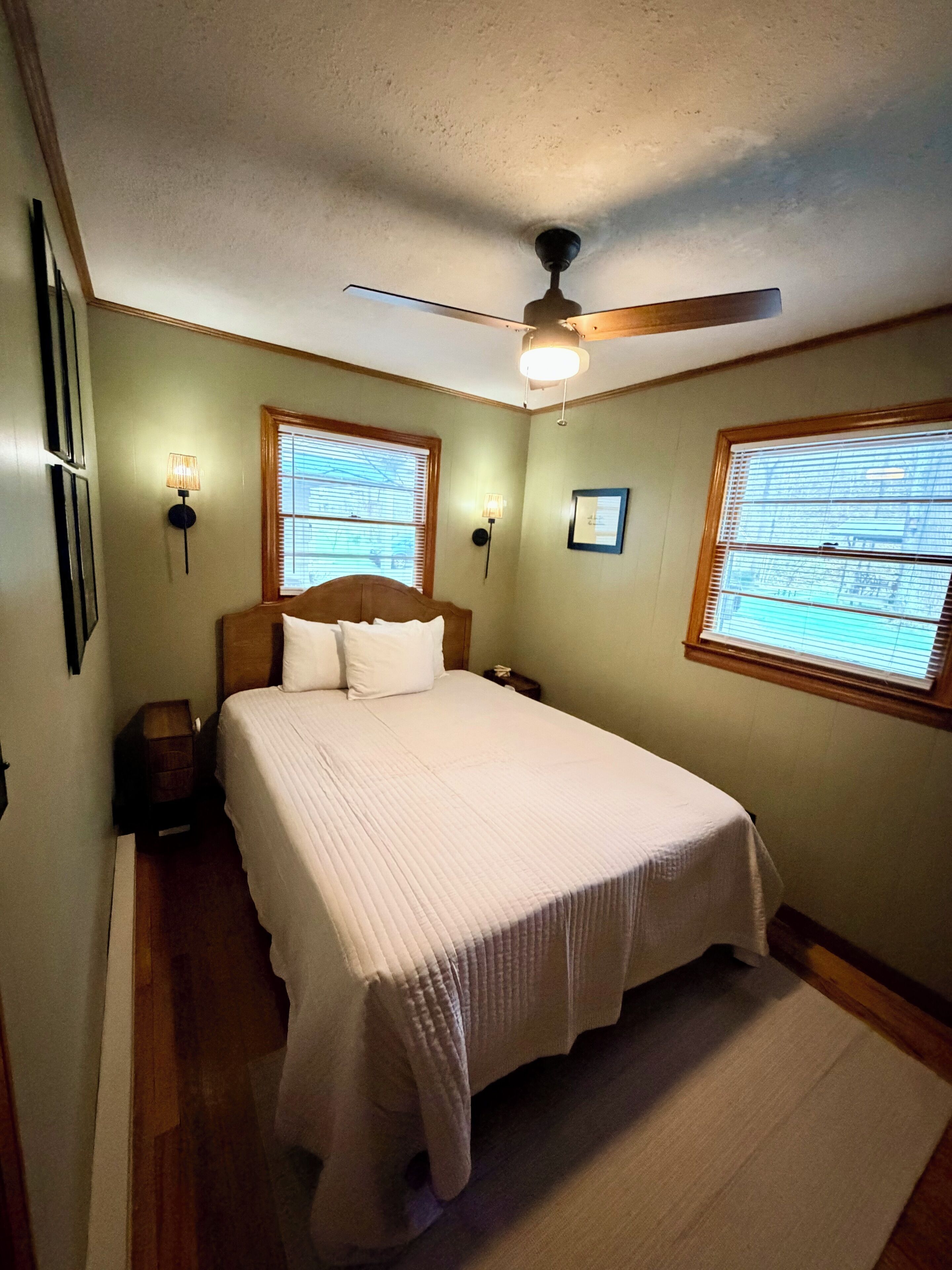 A bedroom with a bed, two wall-mounted lamps, a ceiling fan, and two windows with blinds.