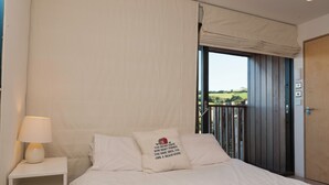 Une chambre à coucher avec un lit, une table de chevet avec une lampe, un oreiller orné d’une citation, une vue sur l’extérieur et une porte en bois.