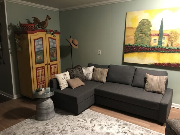 A living room with a dark grey sectional sofa, a patterned rug, a wooden cabinet with decorative items, and a large landscape painting on the wall.