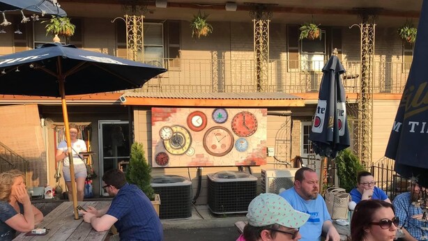 Un espacio al aire libre con mesas y sillas de madera, un mural de relojes en la pared y plantas colgantes.