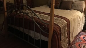 Una cama con dosel de estructura metálica, cabecera de madera y cubrecama estampado. Se aprecia una mesita de noche con lámpara, un cuadro enmarcado en la pared y una ventana con cortinas.