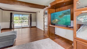 A spacious living room with wooden flooring, a grey sofa, a flat-screen TV mounted on a wooden cabinet, and a large window with a view of greenery. - Gorgeous Colonial House on the Water w/ Pool & Jacuzzi
 (Jacksonville)