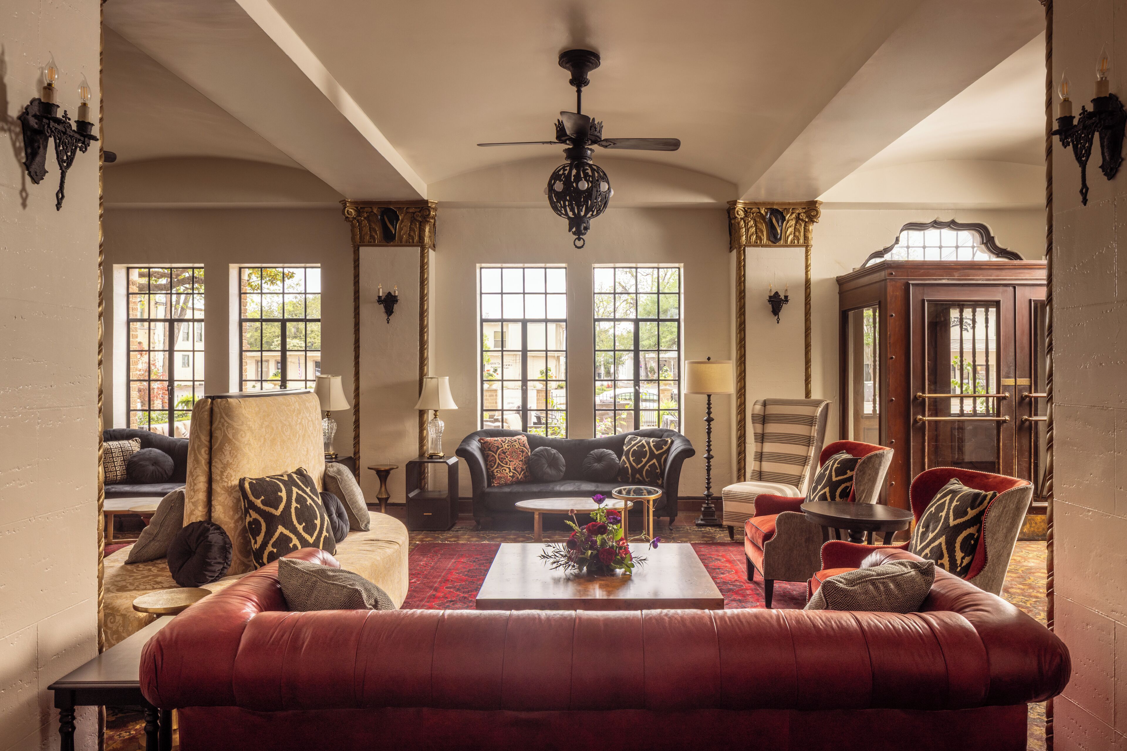 A spacious living room with a red sofa, a black sofa, and a wooden coffee table. There are two windows with views of greenery outside.
