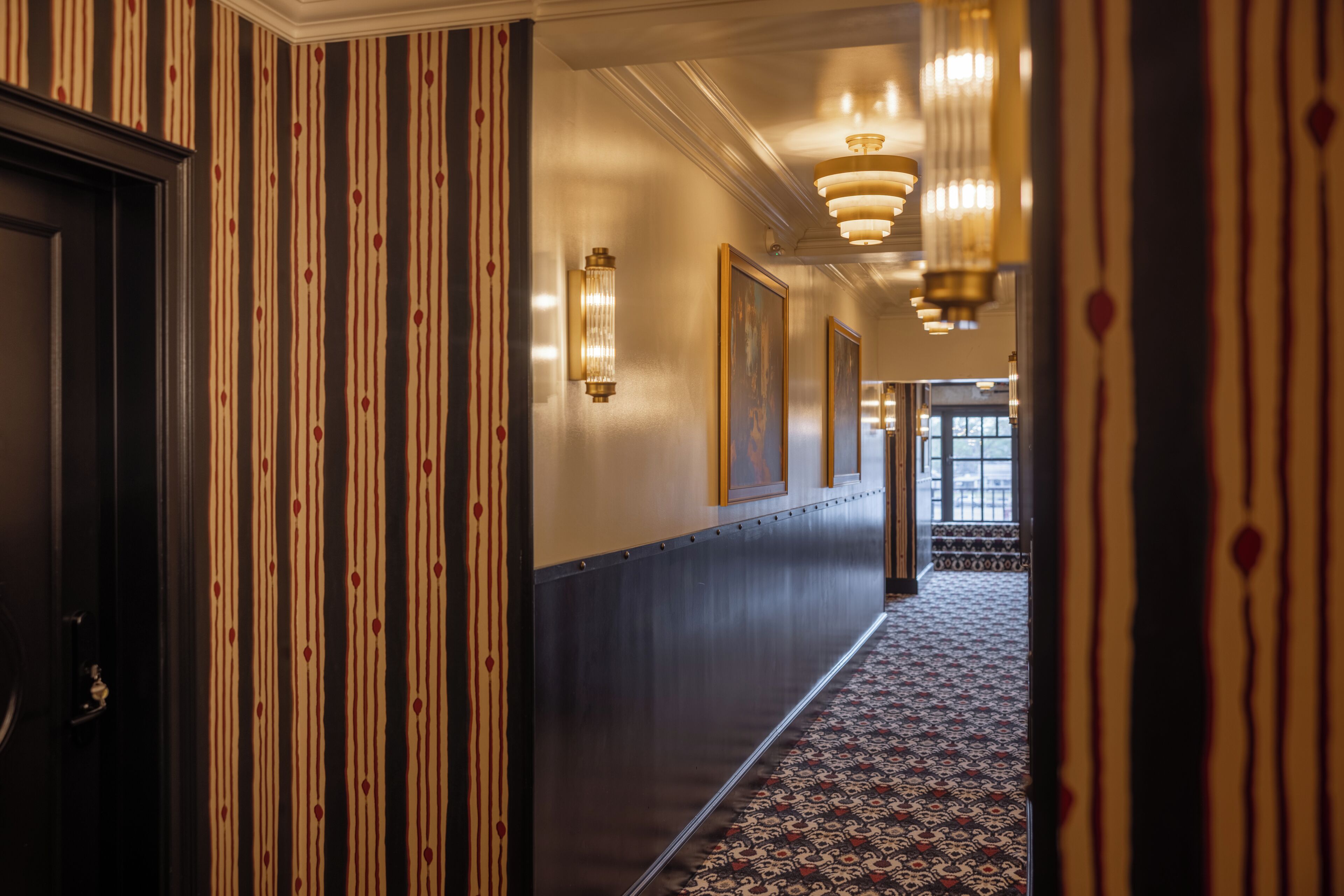 A hotel corridor with striped wallpaper, framed artwork, and patterned carpet.