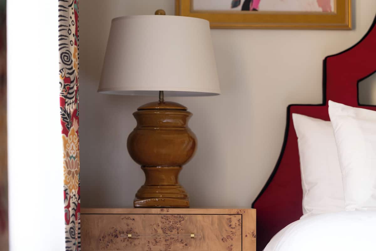Una mesita de noche de madera con una lámpara, una cama con cabecera roja y un cuadro en la pared.
