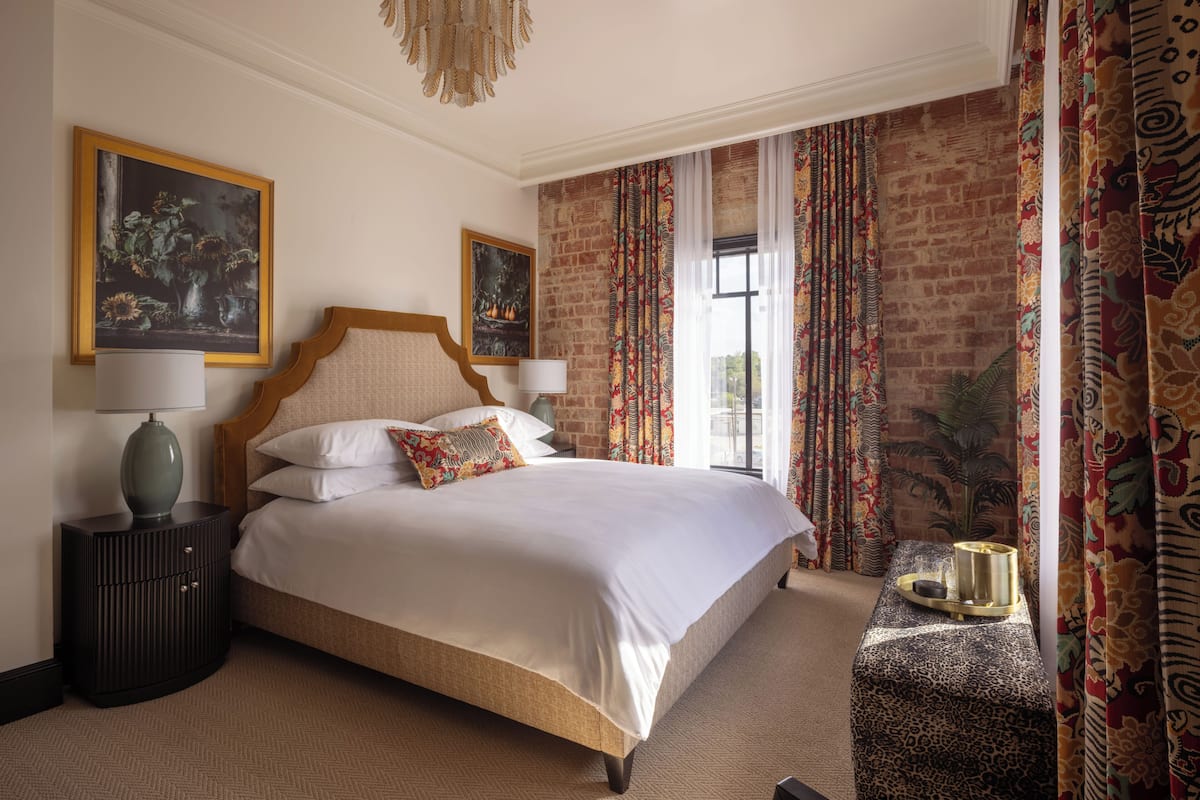 Un dormitorio con una cama grande, una mesita de noche, una lámpara, un cojín decorativo estampado y vistas a un muro de ladrillo.