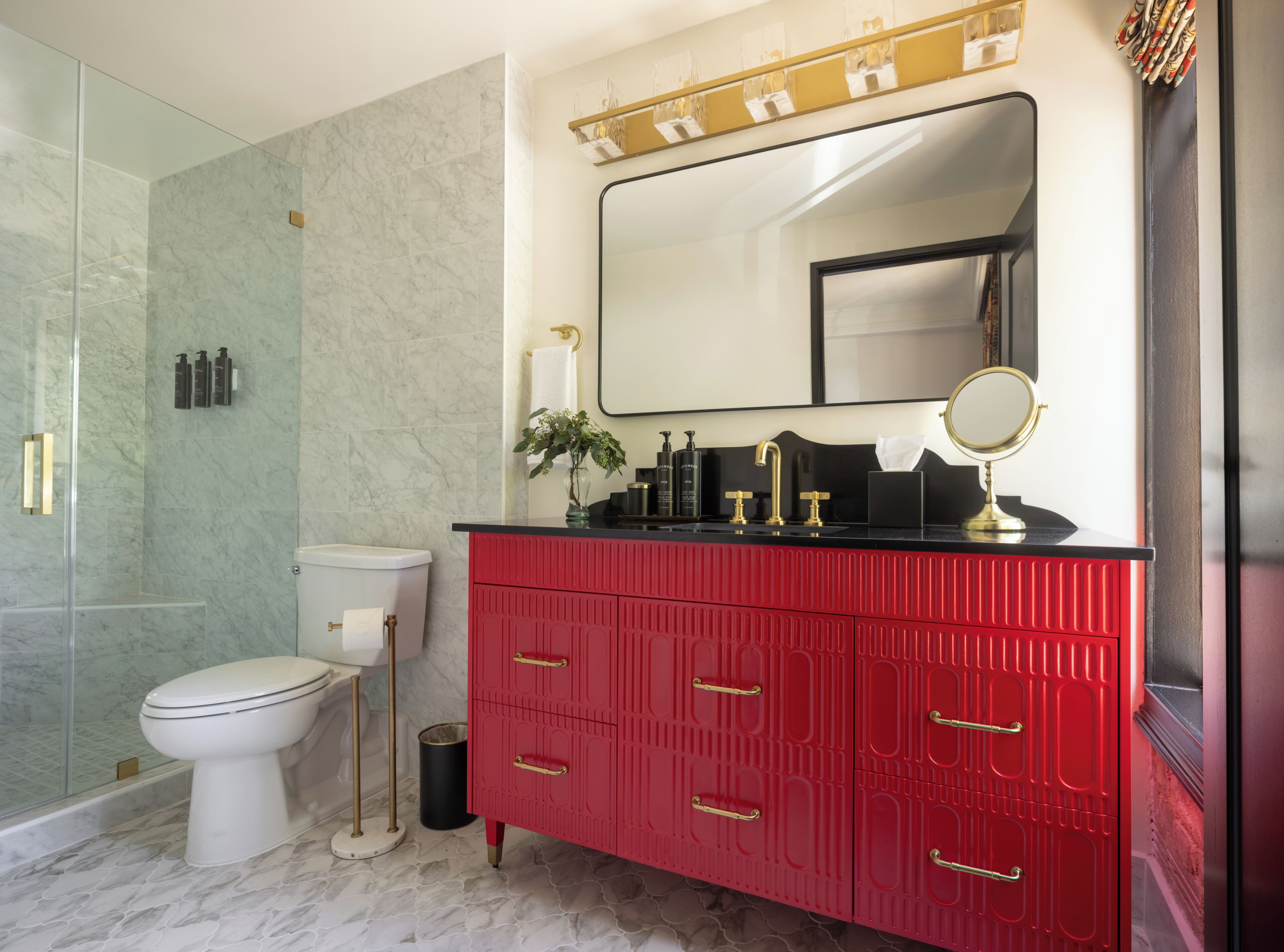 A bathroom with a glass shower enclosure, a white toilet, a red vanity with gold handles, a large mirror, and a black countertop.