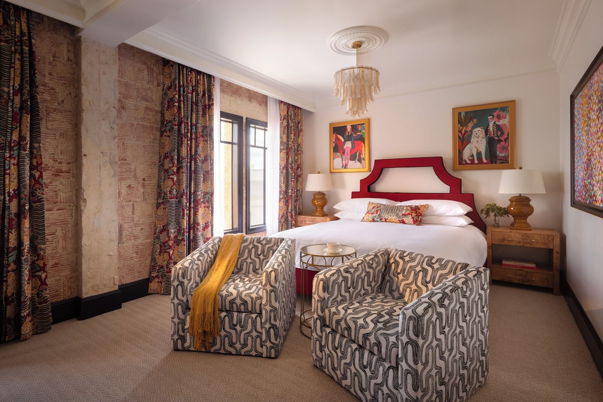 Una habitación de hotel con una cama grande, dos sillones estampados, un candelabro y un cuadro en la pared.