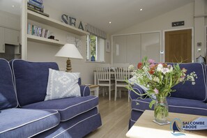 Uma sala de estar com um sofá azul, uma mesa branca com um vaso de flores e uma estante de livros com livros e objetos decorativos.