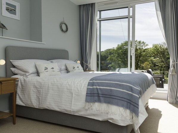 A bedroom with a large bed, a nightstand, a wall-mounted light, a window with curtains, and a view of greenery outside.