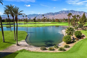 A golf course with a pond, palm trees, and mountains in the background.