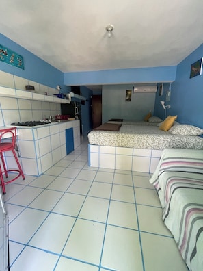 Une pièce compacte avec une kitchenette, un lit recouvert d’une literie à motifs, une chaise rouge et un ventilateur de plafond.