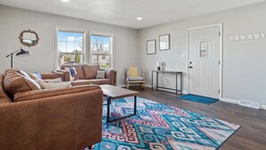 A spacious living room with a brown leather sofa, a coffee table, and a colorful rug. There are two windows providing natural light, a door, and a small table with a lamp. - Gazebo-4BR Ping Pong-Arcade Room (Rapid City)