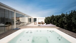 A modern outdoor hot tub area with a wooden deck, glass walls, and a view of a building and trees.