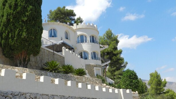 Un edifici blanc de diverses plantes amb finestres i balcons arquejats, envoltat de vegetació i una paret de pedra.