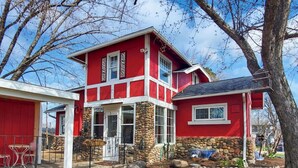 A two-story house with a red exterior and stone accents, featuring a front porch with a table and chairs, and a stone wall in the front yard.