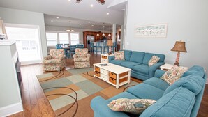 A spacious living room with a blue sofa, floral armchairs, and a coffee table. There is a kitchen in the background with wooden cabinets and a dining area with blue chairs. - Oceanside, Pool Table, Private Pool, Close to Beach! OS9Q, Someday Came (Corolla)