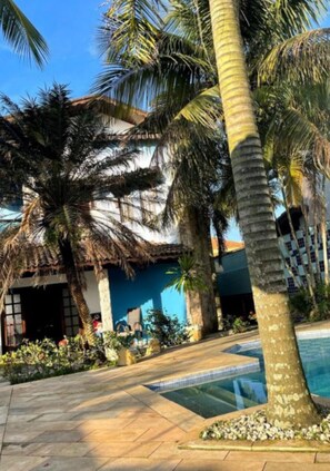 Un espace aménagé au bord de la piscine, avec des palmiers, un bâtiment à toit de chaume et un ciel d’un bleu limpide.