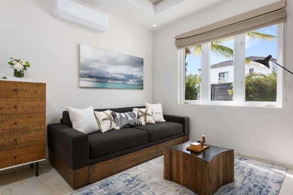 A modern living room with a dark sofa, a wooden cabinet, a coffee table, and a large window with a view of a building and palm trees.