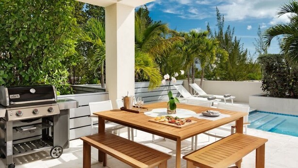 An outdoor patio with a wooden table, benches, a grill, and a pool in the background. - Salt Air and Second Wind Villas (Leeward)
