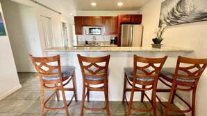 A kitchen with wooden cabinets, a stainless steel refrigerator, and a marble countertop. There are four wooden chairs with cushioned seats. - A Gorgeous Oceanfront Condo! (2BR) (Kapaʻa)