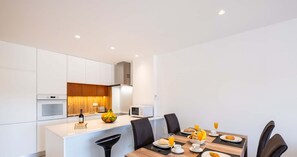 Une cuisine moderne avec des meubles blancs, un îlot central et un coin repas comprenant une table en bois et des chaises noires.