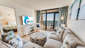 A spacious living room with a large sofa, ottoman, and a television displaying 'BIG FISH Rentals'. A balcony with a view of the ocean is visible. - Coastal condo with beach access & pools (North Myrtle Beach)