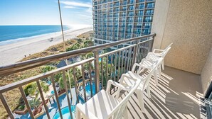 Uma varanda com cadeiras de plástico branco, piscina e vista para a praia e oceano.