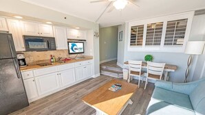 Cozinha moderna com armários brancos, micro-ondas e uma TV exibindo 'BIG FISH'. Há uma mesa de madeira e um sofá azul claro na sala de estar.