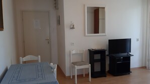 Una habitación con piso de madera, una puerta y una silla blancos, una mesa pequeña con mantel azul, un mueble de TV negro con televisor, y un espejo en la pared.