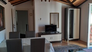 Una habitación con una mesa de comedor, sillas, un televisor sobre un mueble bajo, un armario y un espejo en la pared.