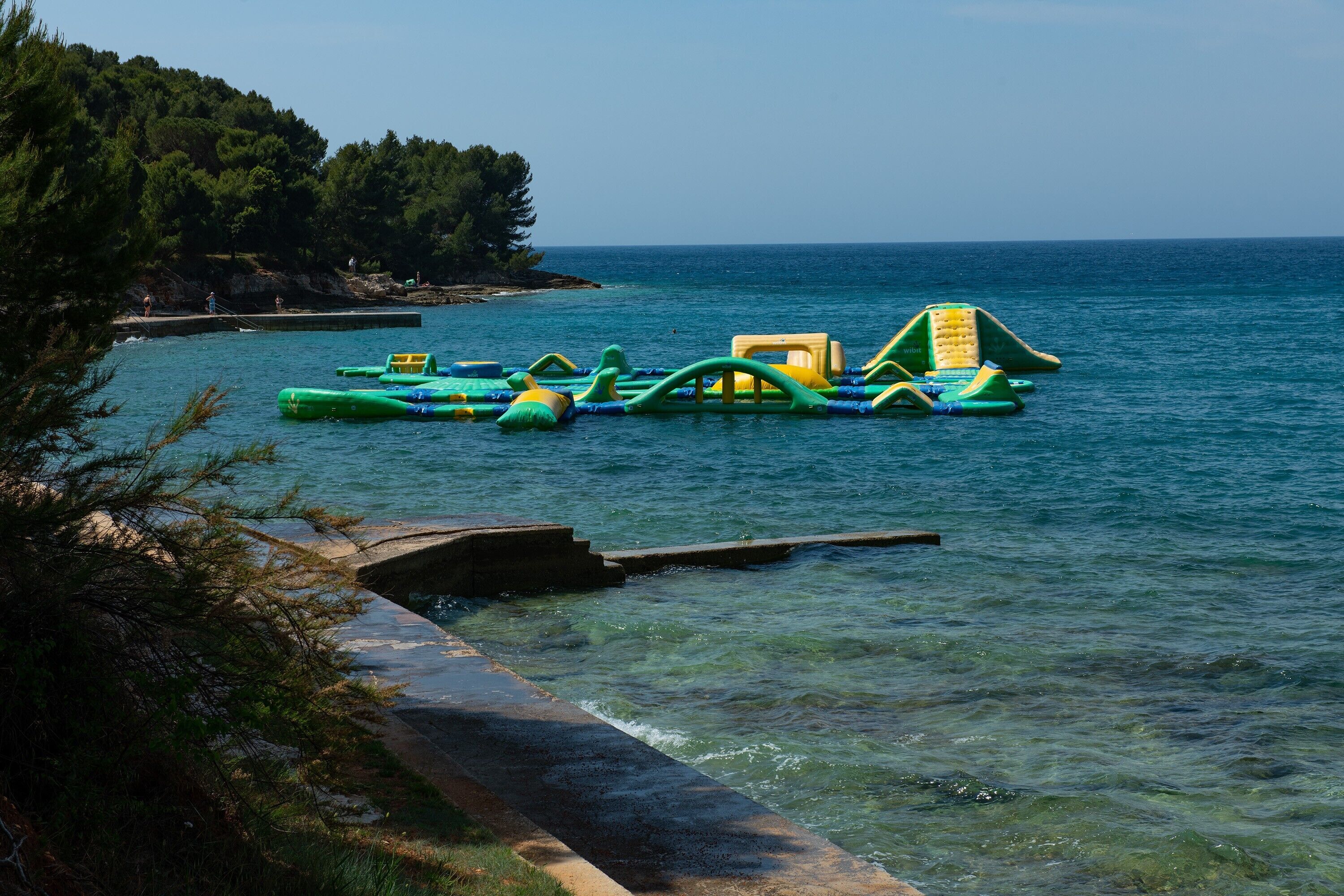 Une zone côtière où l’on trouve des jeux d’eau gonflables sur l’eau et une promenade en béton le long de la plage.