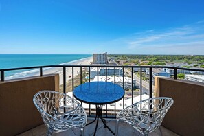 A balcony with a clear view of the beach and ocean, two white chairs, and a round table.