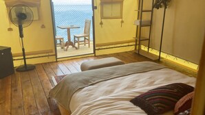 Une chambre avec un plancher en bois, un lit avec un oreiller à rayures, un ventilateur sur pied et une vue sur la mer par une porte ouverte.